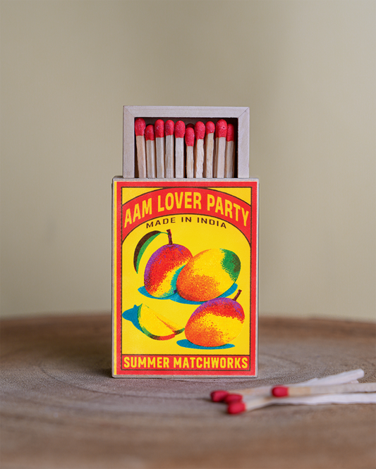 A vintage-style matchbox with a bright yellow cover featuring mango illustrations and the text 'AAM LOVER PARTY' and 'SUMMER MATCHWORKS'. The open box contains red-tipped matches, standing on a wooden surface with a few loose matches in front against a soft beige background.
