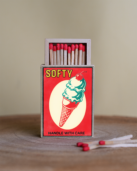 A vintage-style matchbox with the brand name SOFTY on the front, featuring a red cover with a green ice cream cone illustration and the words 'HANDLE WITH CARE.' The matchbox is open, showing wooden matches with red tips, and a few loose matches sit in front on a wooden surface against a soft beige background.